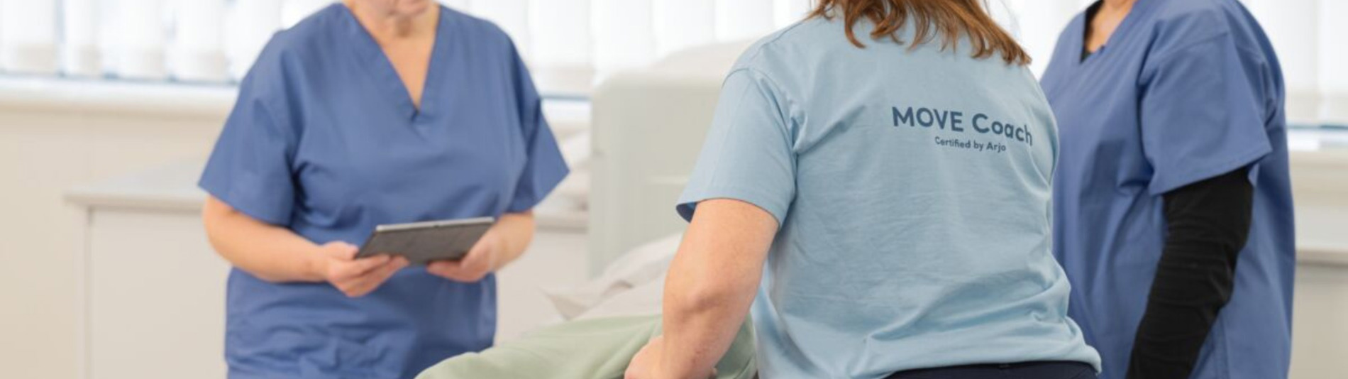 Trois professionnels de la santé dans un cadre clinique, dont l'un porte un t-shirt avec l'inscription "MOVE Coach Certified by Arjo" aidant un patient.
