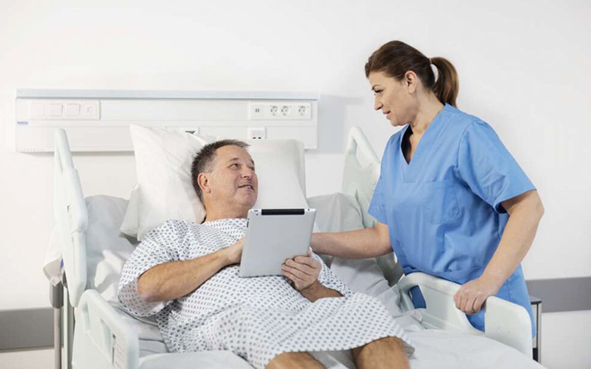 Patient in hospital bed receiving assistance from a nurse using a digital tablet for healthcare support.