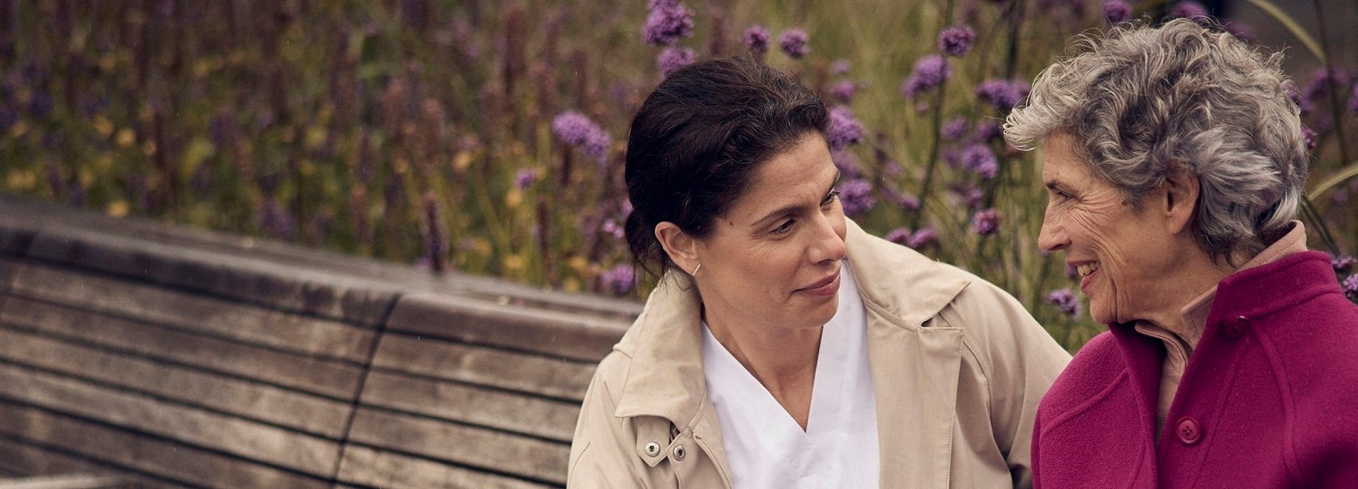 Two individuals seated on a wooden bench in a serene garden with purple flowers in the background, suggesting a calm and natural environment.