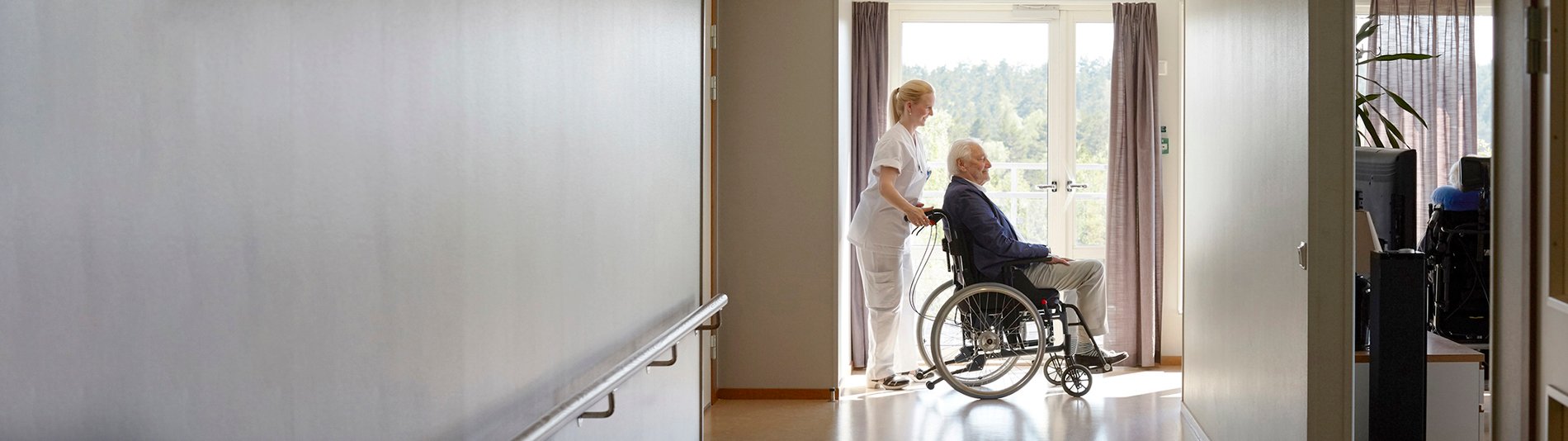 Pflegerin schiebt Rollstuhl über Gang am Fenster